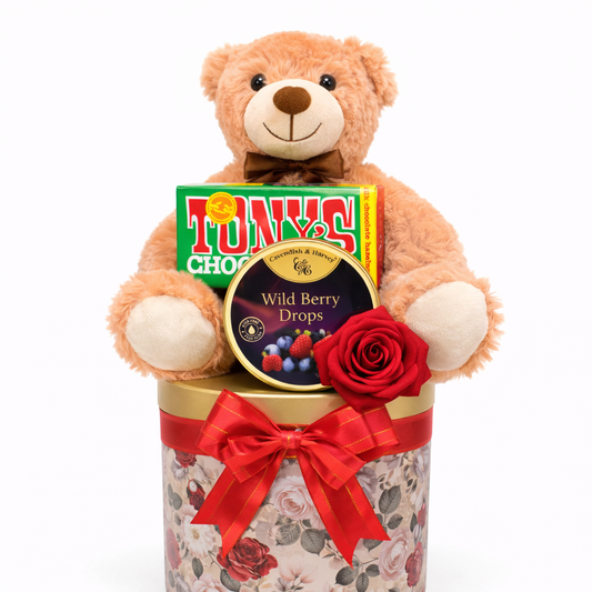 Teddy bear holding a box of Tony's Chocolonely Wild Berry Drops with a floral container on a white background